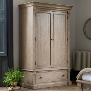 The CHOICEWARDROBE Valletta Wooden Wardrobe features 2 doors, 1 deep drawer, and antique handles in a natural oak finish with a classic panelled design. It stands beside a potted plant, near a radiator and part of a bed.