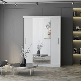 A modern room with a CHOICEWARDROBE Vetra 2 Door Sliding Mirror Wardrobe (150cm, matt white, shelves & hanging rail), a round silver coffee table with a vase of branches, gray walls, built-in shelves, and artwork reflected in the mirror.