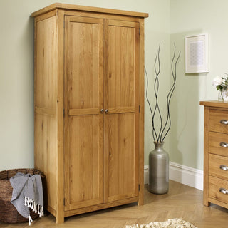 The CHOICEWARDROBE Webworms 2 Door Solid Oak Wardrobe in light oak stands by a green wall, near a silver vase with branches. A matching dresser and a woven basket with a blanket complete the cozy bedroom scene.