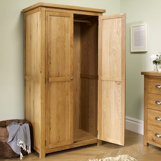 The CHOICEWARDROBE Webworms 2 Door Solid Oak Wardrobe—compact and full hanging in a light natural oak finish—stands open on a wooden floor, beside a chest of drawers and a woven basket holding a blanket.