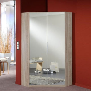 The GEMA 2 Door Corner Wardrobe in oak with white gloss doors stands against red walls, its mirrored finish reflecting a sunlit room with a bench, shoes, a shopping bag, and dried decorative grass.