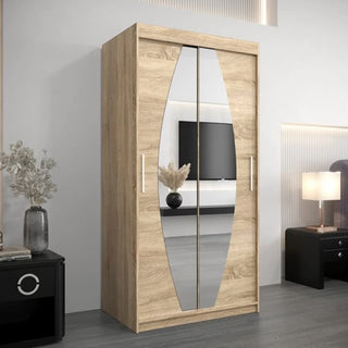 The Eden Mirrored Wardrobe 2 Sliding Doors 100cm in Sonoma Oak stands in a modern room, reflecting a TV. It features light wood tones, vertical handles, and is placed near black furniture topped with a vase and lamp.