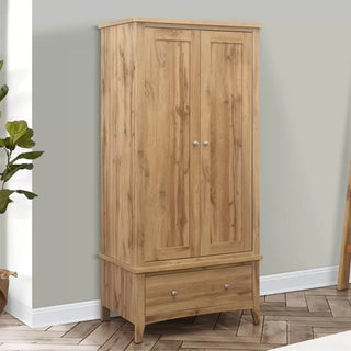 The Hebron Wooden Wardrobe With 2 Doors 1 Drawer In Oak, accented with metal knobs, stands against a light gray wall on a herringbone floor. A plant on the left adds elegance to your bedroom storage solution.