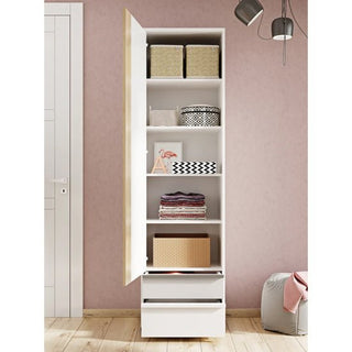 The Indio Kids Wooden Wardrobe With 1 Door in matt white stands against a pink wall, shown with the door open to display baskets, folded towels, boxes, and decorative items on its shelves. Pendant lights and a closed door are nearby.