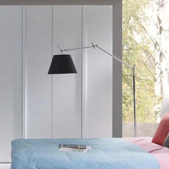 A modern bedroom features a bed with a blue and pink blanket, pillows, a tall floor lamp with a black shade, and an Iowa High Gloss Wardrobe With 4 Hinged Doors in white. A window reveals trees outside.