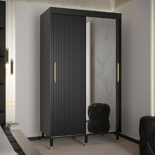 The Millwall 120cm Mirrored Wardrobe With 2 Sliding Doors In Black features gold handles and legs, one ribbed door, and one full-length mirrored door. It stands beside a black pouf on a light-colored floor.