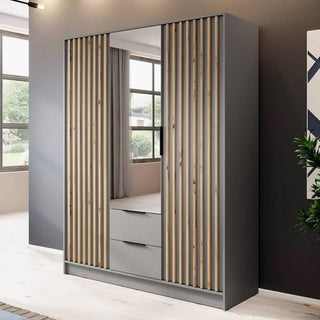 The Newberry Mirrored Wardrobe with 3 hinged doors in grey, featuring vertical wooden slats and two bottom drawers, stands against a dark wall in a brightly lit room with large windows.