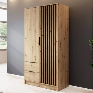 The Newberry Wooden Wardrobe With 2 Hinged Doors 105cm in Artisan Oak, featuring black vertical slats on one door and two lower drawers, stands against a gray wall lit by natural light from a nearby window.