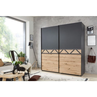 A modern bedroom with the ANDORRA 2 Door Wardrobe (Graphite and Oak, 180cm), featuring sleek sliding doors. The room also has a window, cushioned chair, lamp, and a small table with books and decor for stylish storage.