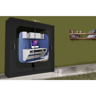 The COVINA 2 Door Mirrored Wardrobe in black (150cm) stands against an olive-green wall, its glossy doors reflecting a modern white desk setup. Nearby, a floating shelf displays books, enhancing this stylish bedroom storage solution.