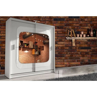 The COVINA 2 Door Mirrored Wardrobe in white (150cm) stands against a brick wall, with glass panels reflecting interior shelves and lights. Above and to the right, a shelf with books and copper pots adds stylish bedroom storage.