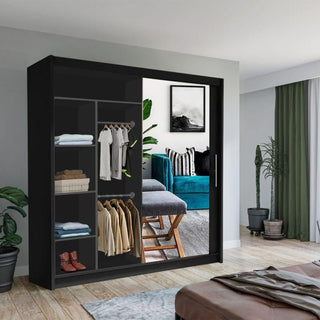 The KEYNES 2 Door Mirrored Wardrobe in black (150cm) is open, displaying neatly arranged clothes, shoes, and boxes. The mirrored doors reflect a living room with a blue sofa, plants, and framed art on the wall.