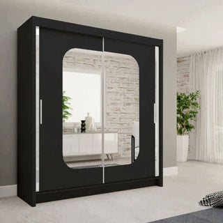 The MERICA 2 Door Mirrored Wardrobe in black (180cm) stands against a light wall in a bright, minimalist bedroom. Its large central mirror reflects a plant and dresser, adding depth and stylish storage to the space.