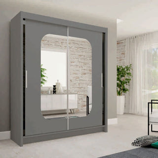 The MERICA 2 Door Mirrored Wardrobe in grey (180cm) stands in a bright, minimalist room with light walls, a plant, and modern furniture. Its mirrored doors reflect a white sideboard and stylish bedroom storage items.