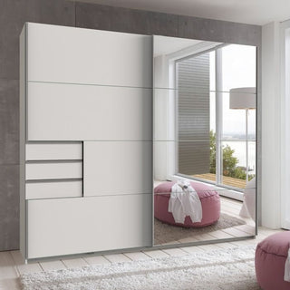 The SOGO 2 Door Mirrored Wardrobe in white (225cm) features sliding doors, three drawers, and multiple panels for storage. It stands in a bright room with pink poufs, a white lamp, and large windows overlooking the water.