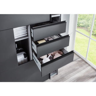 Three partially open black drawers in the SOGO 3 Door Mirrored Wardrobe Oak with Black (270cm) reveal various items inside. The modern wardrobe sits by a large window overlooking green grass, with a wood-patterned floor beneath.