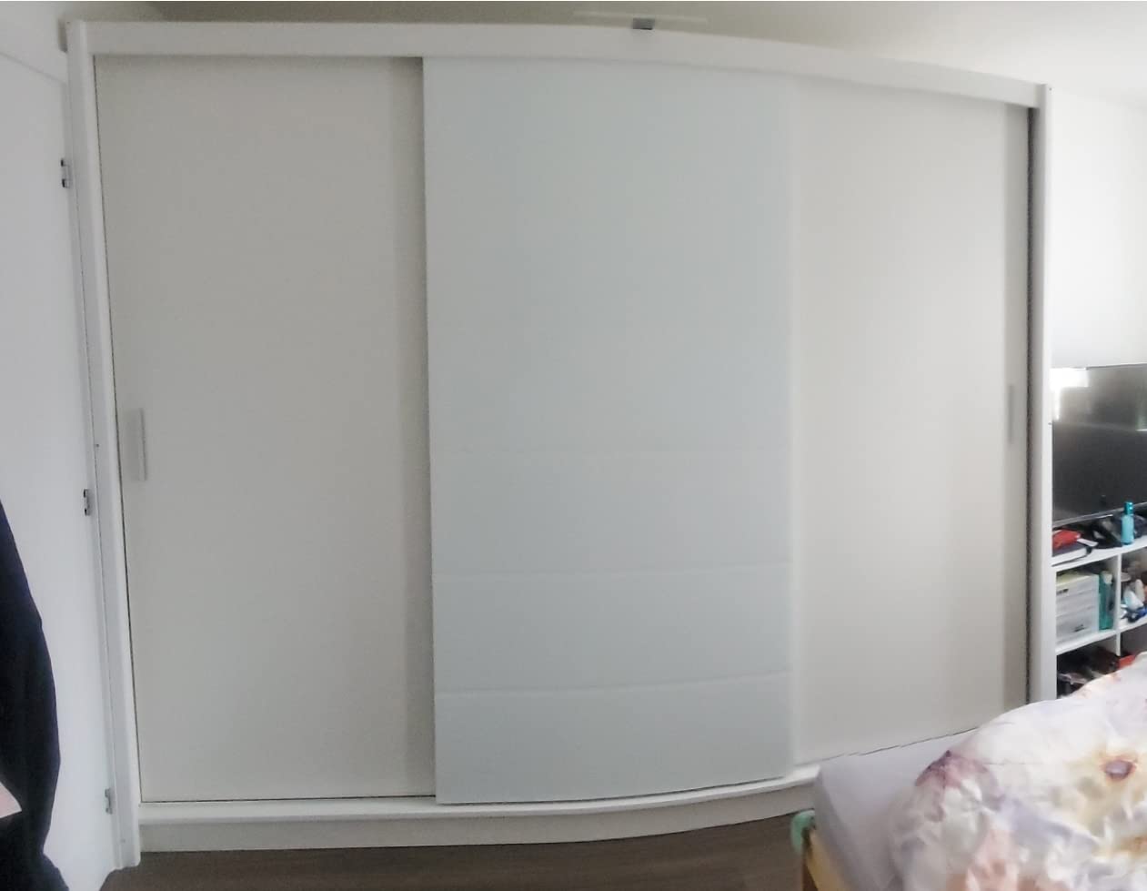 The ROTHERHAM 3 Door Wardrobe White 250cm is closed with sleek sliding doors, concealing a high gloss built-in unit in a room with wooden flooring; part of a bed and shelving unit is visible to the right.