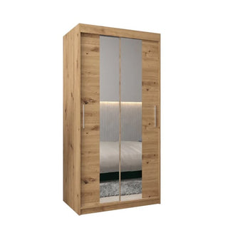 The Tavira I Mirrored Wardrobe, featuring 2 sliding doors in artisan oak, stands in front of a bed and a wall with horizontal paneling and soft lighting.