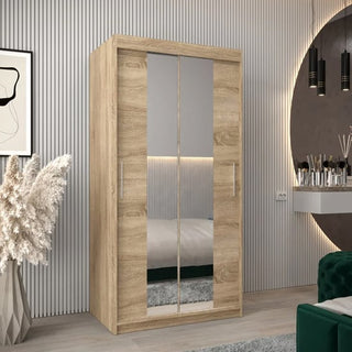 The Tavira I Mirrored Wardrobe with 2 sliding doors in Sonoma Oak (100cm) is showcased in a modern bedroom with a decorative vase of pampas grass, a green upholstered chair, and a round mirror above a floating vanity.