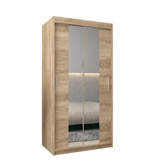 The Tavira I Mirrored Wardrobe, 100cm in Sonoma Oak, features two sliding doors with vertical metal handles and stands against a plain white background. Its mirrors reflect a bed and part of a wall.