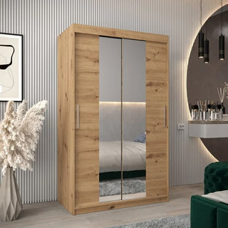 The Tavira I Mirrored Wardrobe with 2 sliding doors in Artisan Oak stands in a modern bedroom, alongside a green velvet chair, decorative vase with pampas grass, round mirror, and wall art.
