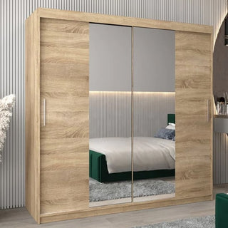 The Tavira I Mirrored Wardrobe with 2 sliding doors in Sonoma Oak (200cm) stands in a modern bedroom, paired with a bed, grey rug, and a desk holding cosmetics in the background.