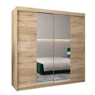 The Tavira I Mirrored Wardrobe with 2 sliding doors (200cm) in Sonoma Oak features mirrored panels and metallic handles, reflecting a modern bedroom with a striped wall, green bed, pillows, and a gray rug.