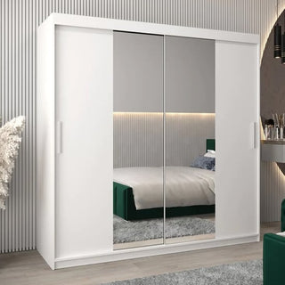 The Tavira I Mirrored Wardrobe with 2 sliding doors (200cm, white) stands in a modern bedroom. Its mirrored panels reflect a bed with green velvet upholstery, a gray rug, and minimalist decor.