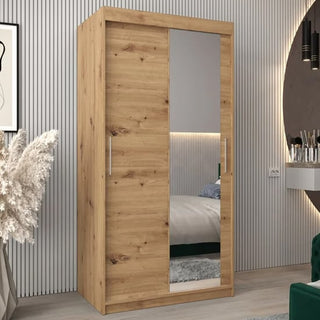 The Tavira II Mirrored Wardrobe 2 Sliding Doors 100cm in Artisan Oak is placed in a modern room with striped walls, decorative plants, and a desk and chair partially visible.