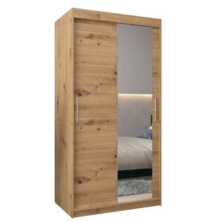 The Tavira II Mirrored Wardrobe features 2 sliding doors, a full-length mirror, and metal handles. Crafted in Artisan Oak with a natural wood grain finish, it measures 100cm wide. A bed and wall are partially visible in the mirror’s reflection.