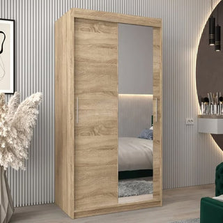 The Tavira II Mirrored Wardrobe with 2 sliding doors in Sonoma Oak (100cm) features a full-length mirror and stands in a modern bedroom with a striped wall, decorative vase, framed artwork, and round vanity mirror.