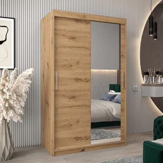 A Tavira II Mirrored Wardrobe with 2 sliding doors (120cm, Artisan Oak) stands in a modern bedroom with gray walls, a large round mirror, pampas grass decor, and a partially visible bed.