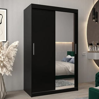 A Tavira II Mirrored Wardrobe with 2 sliding doors (120cm, black) stands in a modern bedroom with vertical striped walls and neutral decor. Its mirrored door reflects part of the bed and bedside area.