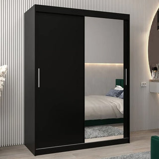 The Tavira II Mirrored Wardrobe 2 Sliding Doors 150cm in black stands against the wall in a modern bedroom, its large mirrored panel reflecting part of the room and bed.