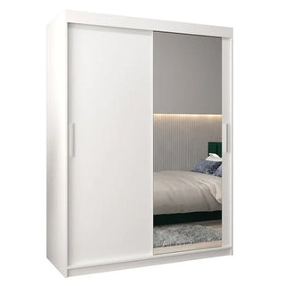 The Tavira II Mirrored Wardrobe with 2 sliding doors (150cm, white) features a mirrored panel that reflects a modern bedroom setting with a bed, gray headboard, pillows, and accent-lit textured wall.