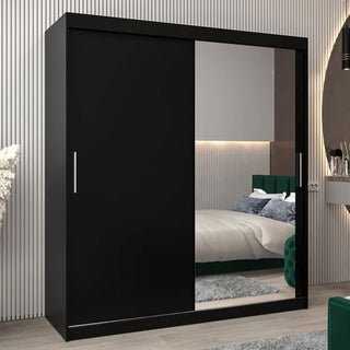 The Tavira II Mirrored Wardrobe 2 Sliding Doors 180cm in Black stands in a modern bedroom with a neatly made bed, green headboard, and light-colored vertical-lined walls.