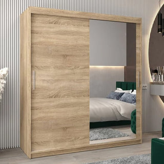 The Tavira II Mirrored Wardrobe with 2 sliding doors (180cm) in Sonoma Oak stands in a modern bedroom, with its mirrored door reflecting a bed, rug, and decorative wall accents for added style and function.
