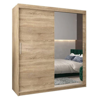 The Tavira II Mirrored Wardrobe (180cm) features 2 sliding doors in Sonoma Oak—one with a natural wood finish and the other a large mirror, reflecting elements of the bedroom such as the bed and rug.
