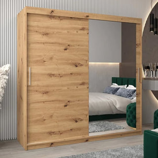 The Tavira II Mirrored Wardrobe (200cm, Artisan Oak) with one solid wood and one mirrored sliding door stands in a modern bedroom featuring a bed, green upholstered headboard, wall paneling, and a makeup table.