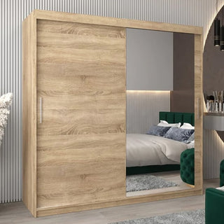 A Tavira II Mirrored Wardrobe with 2 sliding doors (200cm, Sonoma Oak) stands in a modern bedroom with one mirrored door and one wood panel, paired with a green upholstered bed, gray rug, and chic wall decor.
