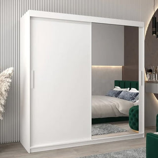 The Tavira II Mirrored Wardrobe with 2 sliding doors in white stands in a modern bedroom, reflecting a green velvet bed, stylish décor, part of a desk, and featuring neutral walls with wooden flooring.
