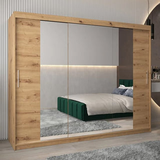 The Tavira II Mirrored Wardrobe with 3 sliding doors in Artisan Oak (250cm) stands in a modern bedroom with a green bed, gray rug, wood flooring, and vertical wall paneling.