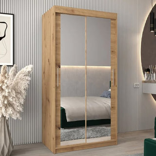 The Tavira III Mirrored Wardrobe with 2 sliding doors in artisan oak stands against a striped wall, its mirrored surface reflecting a bed with green bedding, a gray rug, pampas grass in a vase, and a round wall mirror.