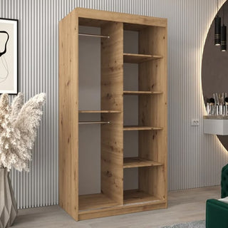 The Tavira III Mirrored Wardrobe with 2 sliding doors (100cm) in artisan oak stands against a striped wall, showcasing its elegant natural finish, while decorative objects and nearby furniture complete the setting.