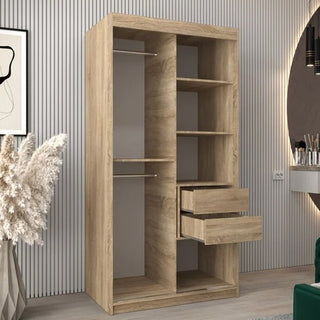 The Tavira III Mirrored Wardrobe with 2 sliding doors (100cm, Sonoma Oak) stands against a striped wall, featuring multiple shelves, two hanging rods, and two partially opened drawers, with a plant and vanity nearby.