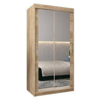 The Tavira III Mirrored Wardrobe with 2 sliding doors in Sonoma Oak (100cm) features a light wood finish, vertical handles, and mirrored panels that reflect the bedroom’s bed, headboard, and gray rug.