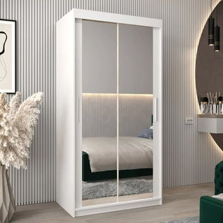 The Tavira III Mirrored Wardrobe (100cm, 2 Sliding Doors, White) stands in a modern bedroom with a bed, vanity, round mirror, pampas grass decor, and vertical-striped wall paneling.