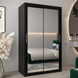 The Tavira III Mirrored Wardrobe with 2 sliding doors in black stands in a modern bedroom with a gray rug, wall art, vanity, pampas grass, vertical paneling, and soft lighting.