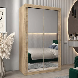 The Tavira III Mirrored Wardrobe with 2 sliding doors in Sonoma Oak (120cm) stands in a modern bedroom with vertical wall panels, a green-headboard bed, a round mirror, and decorative grass in a vase.