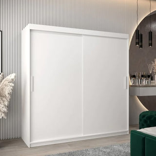 A Tavira Wooden Wardrobe 2 Sliding Doors 200cm In White stands against a light vertical panel wall in a modern room featuring a vanity table, decorative accents, and a partially visible green sofa.